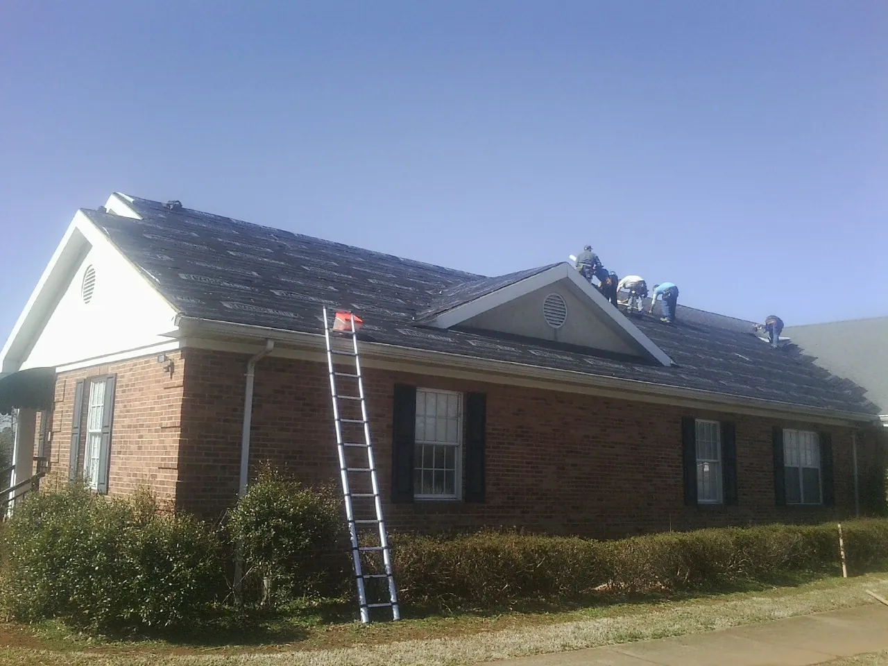 Crew tearing shingles off a brick ranch roof with a ladder set — Emergency Roof Repair work by our crew in New Goshen