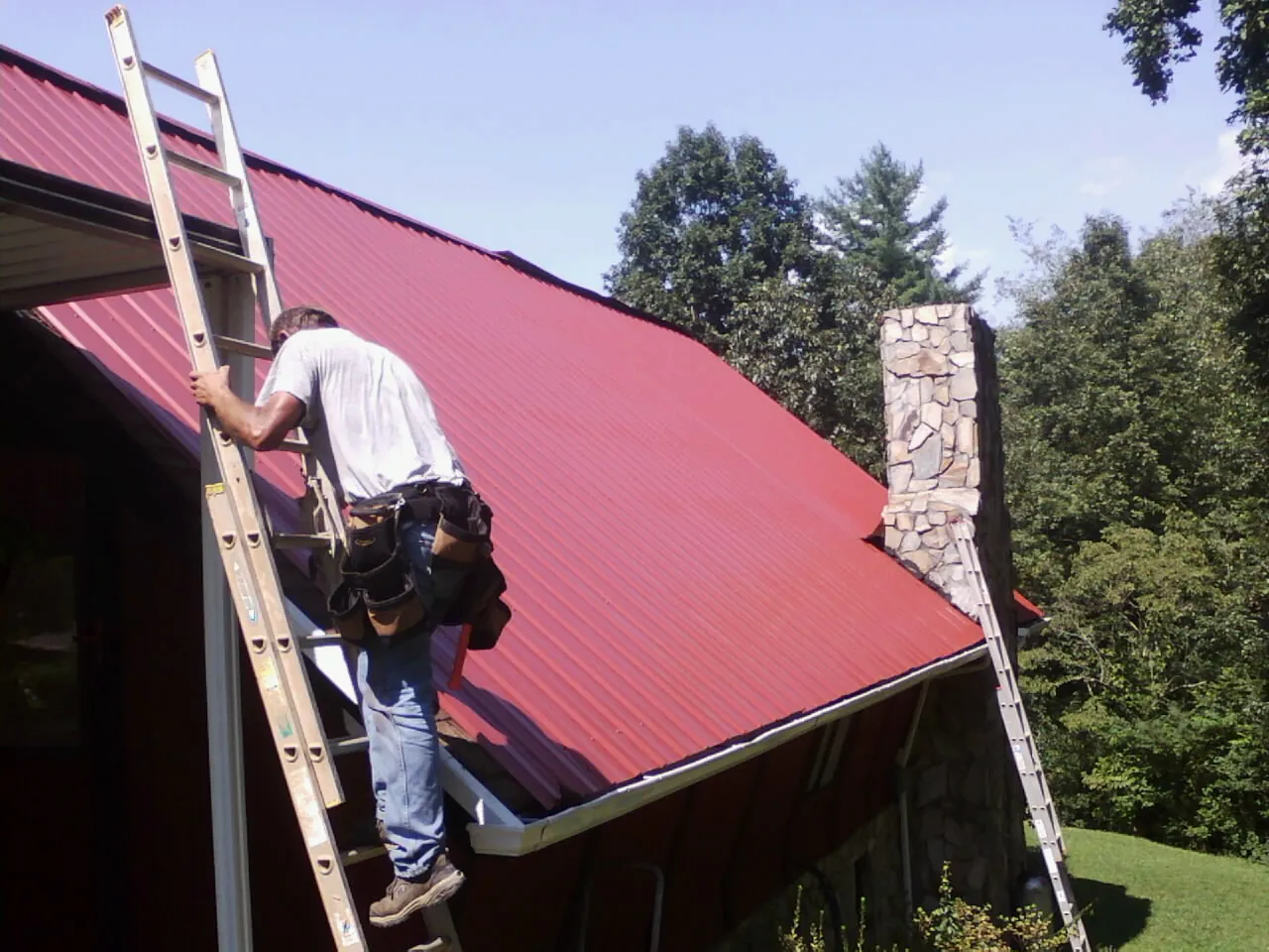 Roofer on a ladder beside a red metal roof and stone chimney — Soffit & Fascia Repair by Roofing Brothers in New Goshen